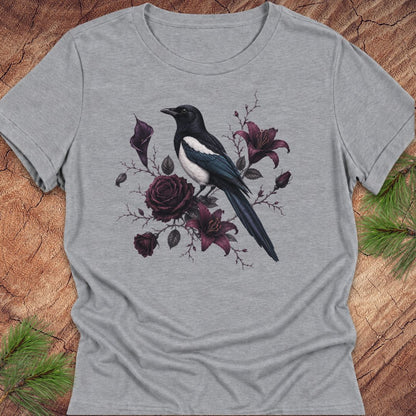 A heather gray short-sleeve crewneck t-shirt featuring a graphic print of a black and white magpie perched among dark purple roses and lilies, displayed flat on a rustic wood background with pine needles.
