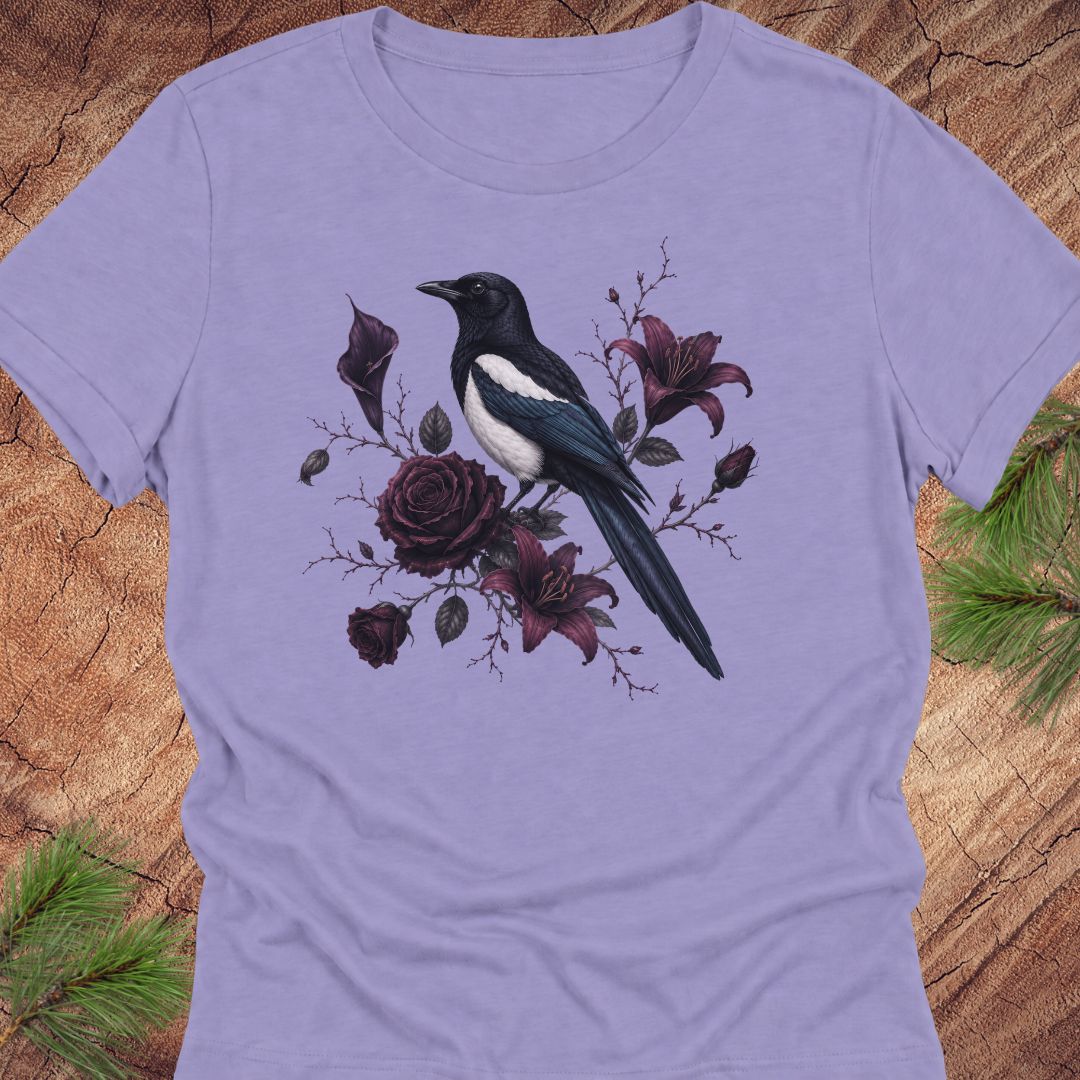 A lavender short-sleeve crewneck t-shirt featuring a graphic print of a black and white magpie perched among dark purple roses and lilies, displayed flat on a rustic wood background with pine needles.