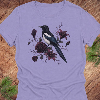 A lavender short-sleeve crewneck t-shirt featuring a graphic print of a black and white magpie perched among dark purple roses and lilies, displayed flat on a rustic wood background with pine needles.