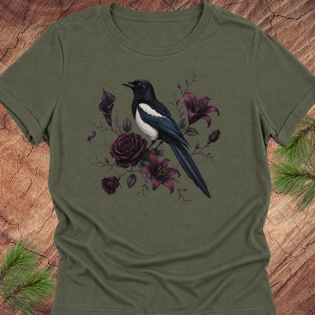 A green short-sleeve crewneck t-shirt featuring a graphic print of a black and white magpie perched among dark purple roses and lilies, displayed flat on a rustic wood background with pine needles.