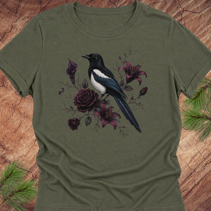A green short-sleeve crewneck t-shirt featuring a graphic print of a black and white magpie perched among dark purple roses and lilies, displayed flat on a rustic wood background with pine needles.