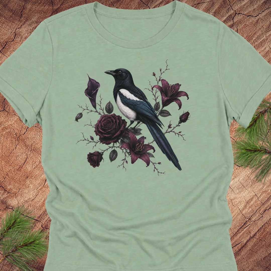 A sage green short-sleeve crewneck t-shirt featuring a graphic print of a black and white magpie perched among dark purple roses and lilies, displayed flat on a rustic wood background with pine needles.