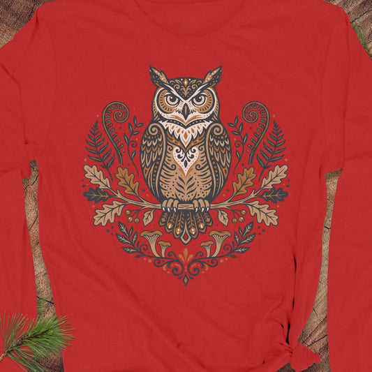 A red long-sleeve t-shirt featuring a detailed graphic of an owl perched on an oak branch with autumn leaves and ferns. The shirt is laid flat on a rustic wood background with a pine sprig in the corner.