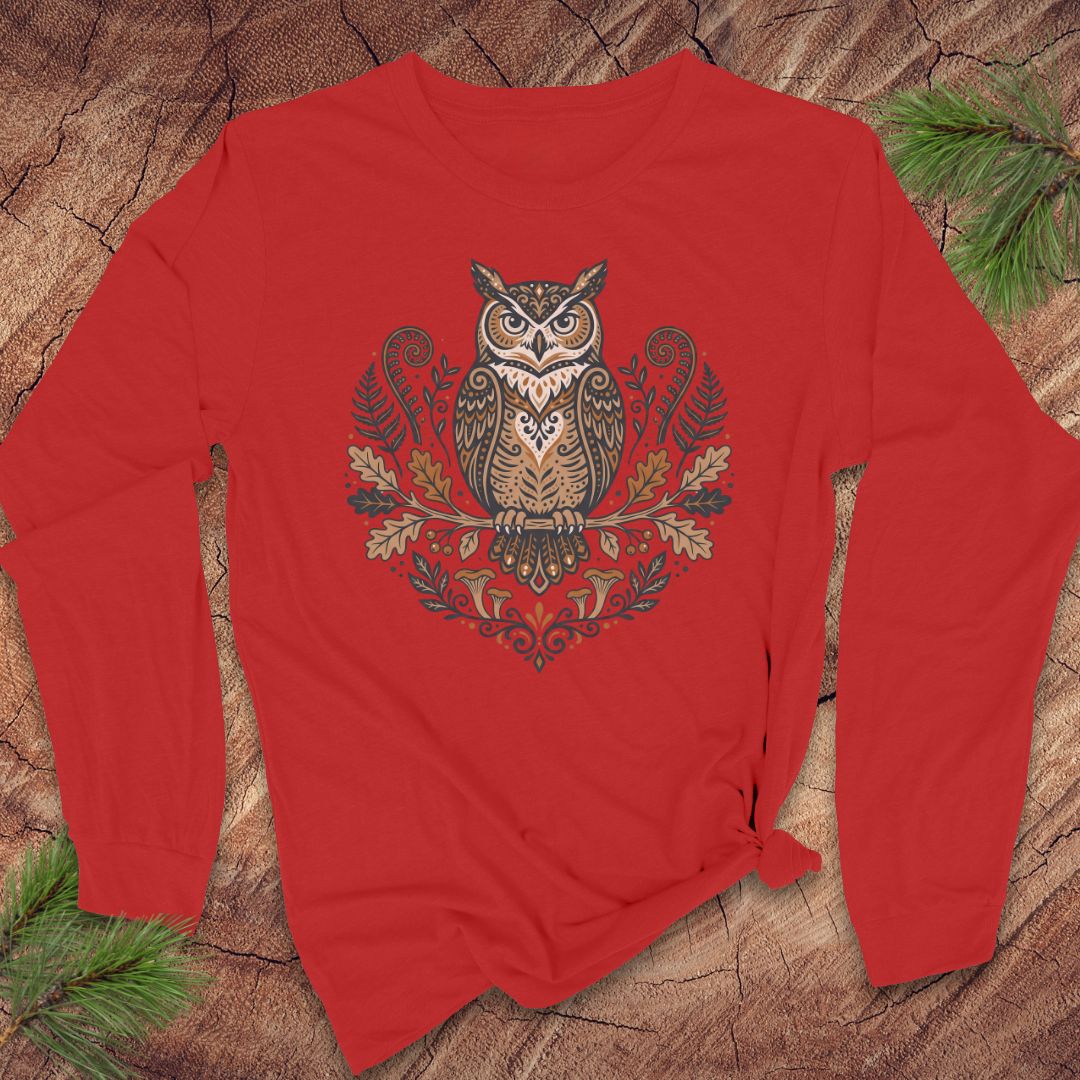A red long-sleeve t-shirt with a detailed owl graphic on the front, styled flat on a rustic wood background with pine needles.