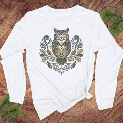 A white long-sleeve t-shirt with a detailed owl graphic on the chest, laid flat on a rustic wood background with pine branches in the corners.