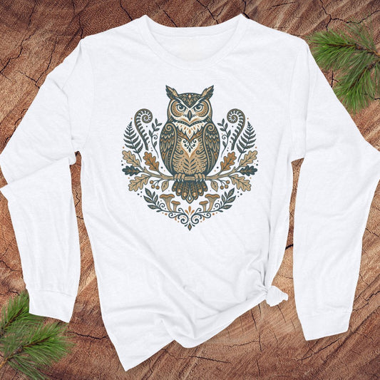 A white long-sleeve t-shirt with a detailed owl graphic on the chest, laid flat on a rustic wood background with pine branches in the corners.