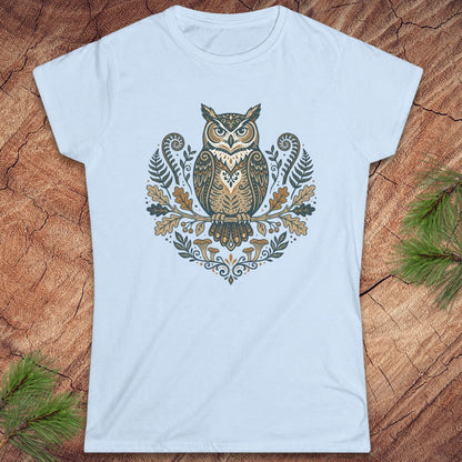 A light blue short-sleeve crewneck t-shirt featuring a detailed graphic of an owl surrounded by ferns, leaves, and mushrooms in earthy tones, laid flat on a rustic wood background with pine branches in the corners.
