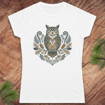 A white short-sleeve crewneck t-shirt featuring a detailed graphic of an owl surrounded by leaves and ferns in earthy brown, beige, and muted green tones. The t-shirt is displayed flat on a textured wood background with pine branches in the corners.