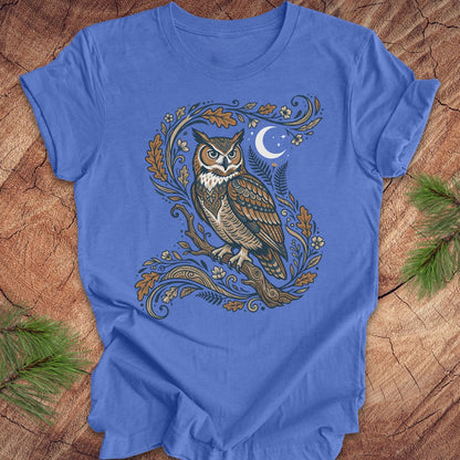 A blue short-sleeve crewneck t-shirt featuring a detailed graphic of a owl perched on a branch with leaves and a crescent moon, laid flat on a rustic wood background with pine branches in the corners.