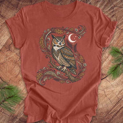 A rust-colored short-sleeve t-shirt with a detailed graphic of an owl perched on a leafy branch under a crescent moon, laid flat on a rustic wood background with pine branches.