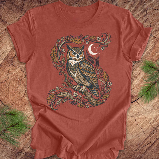 A rust-colored short-sleeve t-shirt with a detailed graphic of an owl perched on a leafy branch under a crescent moon, laid flat on a rustic wood background with pine branches.