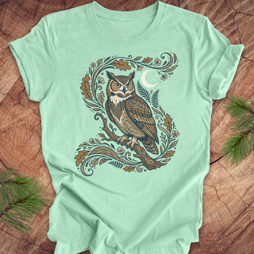 A mint green short-sleeve t-shirt with a detailed graphic of a Great Horned Owl surrounded by autumn leaves, ferns, and a crescent moon, laid flat on a rustic wood background with pine branches.