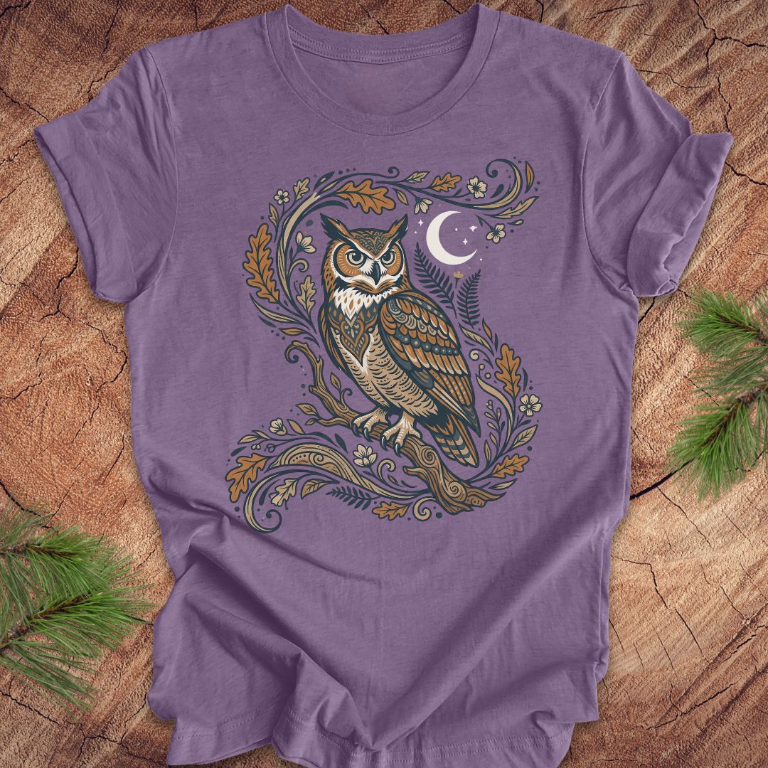 A purple short-sleeve crewneck t-shirt featuring a detailed graphic of a owl perched on a decorative branch with leaves, ferns, and a crescent moon. The t-shirt is laid flat on a rustic wood surface with pine branches visible in the corners.