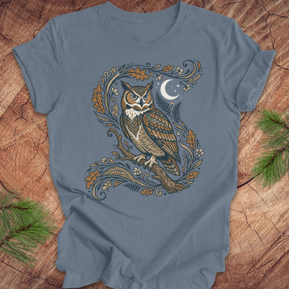 A slate blue short-sleeve t-shirt with a detailed graphic of an owl perched on a decorative branch with a crescent moon, laid flat on a rustic wood background with pine needles in the corners.