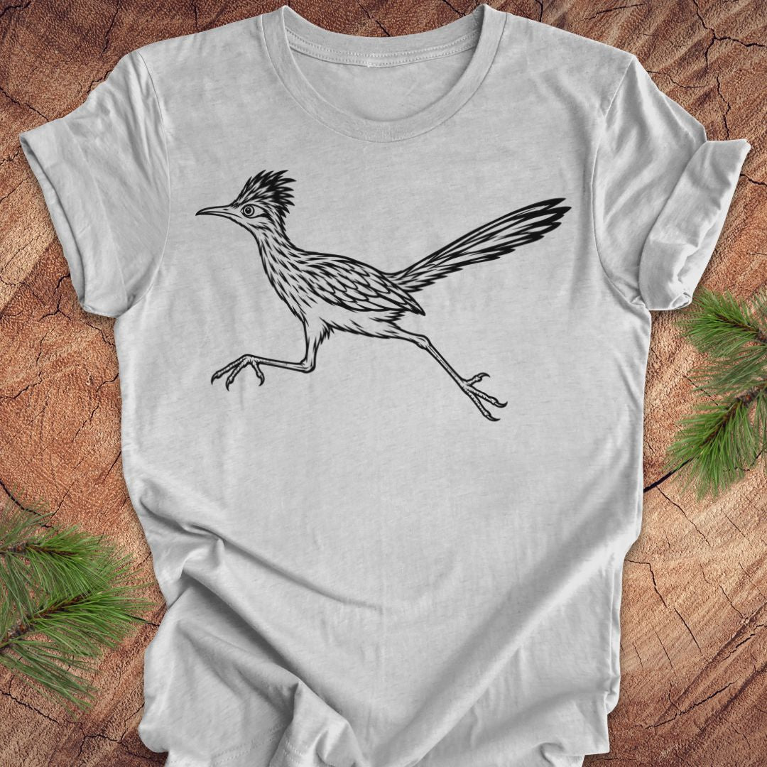 A light gray short-sleeve t-shirt with a black line art illustration of a roadrunner, laid flat on a rustic wood background with pine branches in the corners.