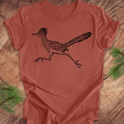 A terracotta-colored short-sleeve t-shirt featuring a black line art illustration of a roadrunner, laid flat on a rustic wood background with pine branches in the corners.