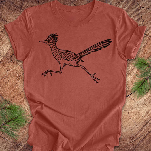 A terracotta-colored short-sleeve t-shirt featuring a black line art illustration of a roadrunner, laid flat on a rustic wood background with pine branches in the corners.