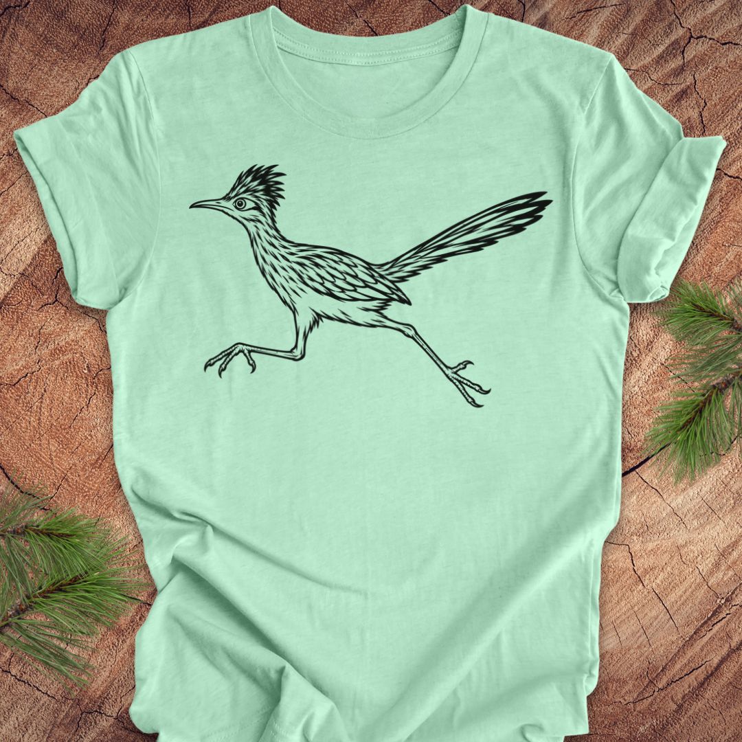 A mint green short-sleeve t-shirt featuring a black line art illustration of a roadrunner, laid flat on a rustic wood background with pine branches in the corners.