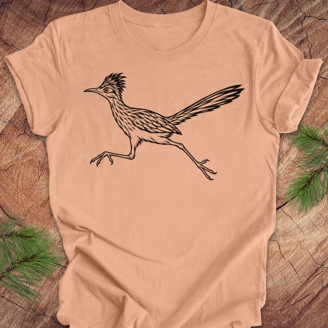 A peach-colored short-sleeve t-shirt with a black line art illustration of a roadrunner, laid flat on a rustic wood background with pine branches in the corners.