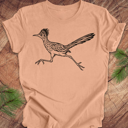 A peach-colored short-sleeve t-shirt with a black line art illustration of a roadrunner, laid flat on a rustic wood background with pine branches in the corners.
