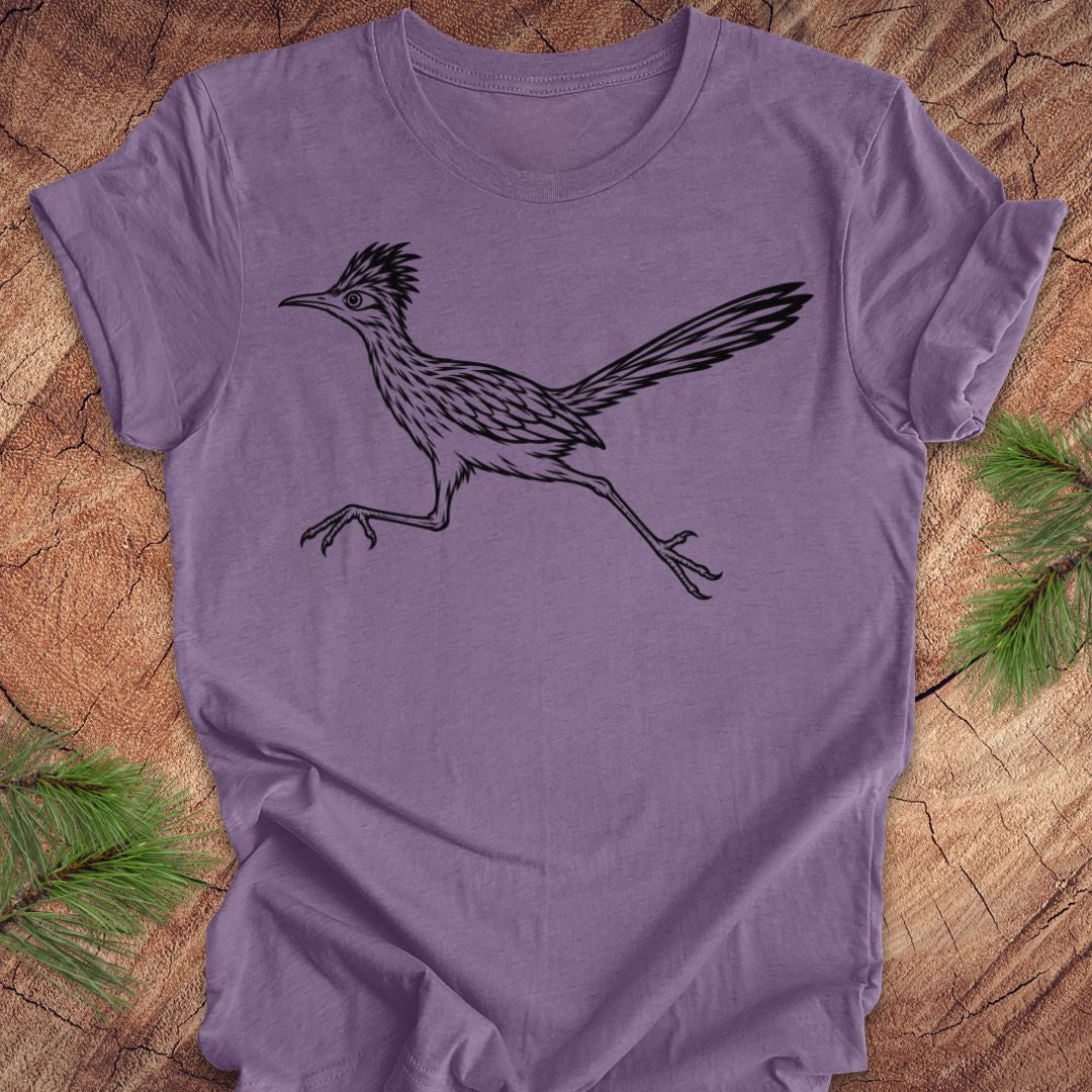 A lavender purple short-sleeve t-shirt featuring a black line art graphic of a roadrunner bird, laid flat on a rustic wood background with pine needles in the corners.
