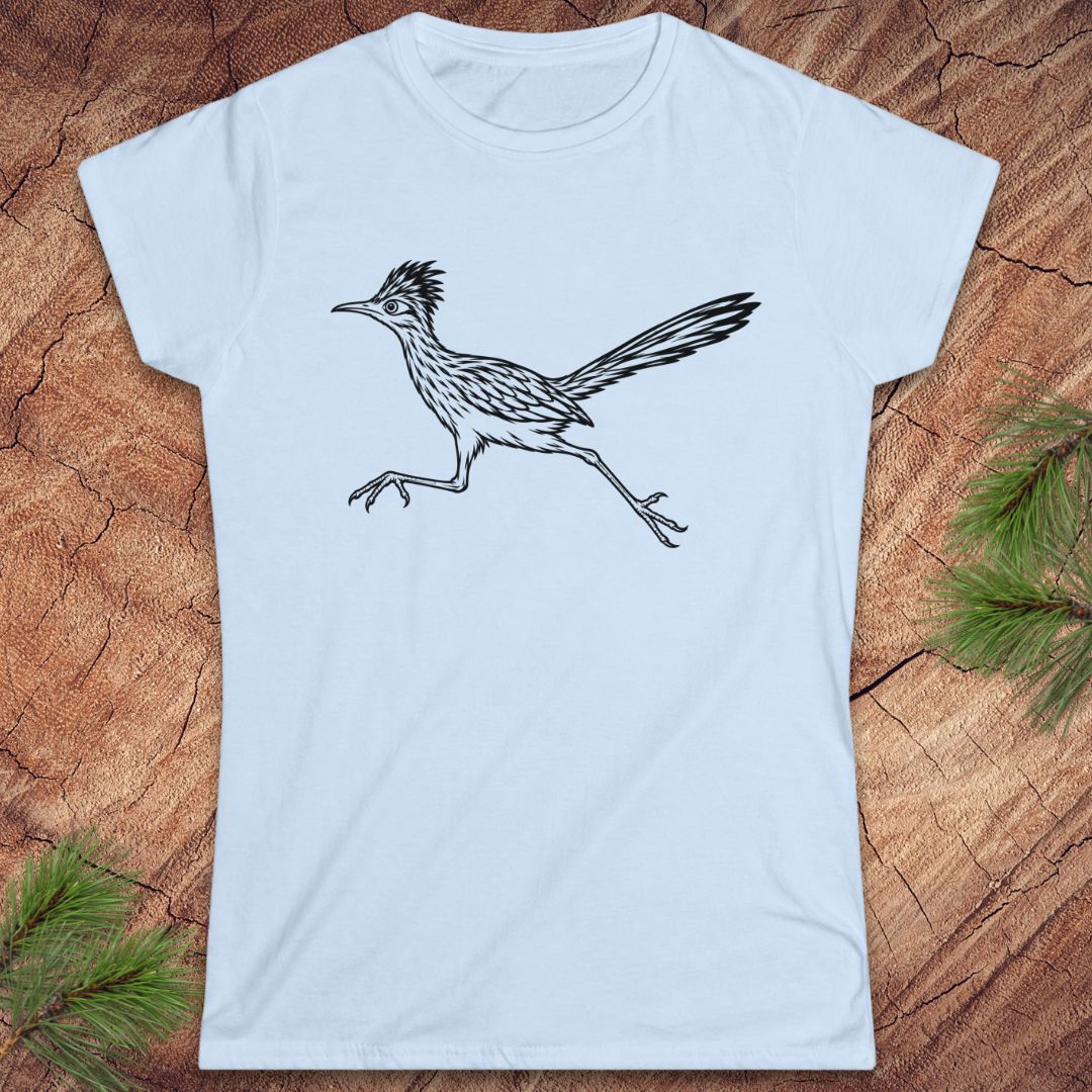 A light blue short-sleeve crewneck t-shirt featuring a black line art illustration of a running roadrunner bird, displayed flat on a rustic wood background with pine needles in the corners.