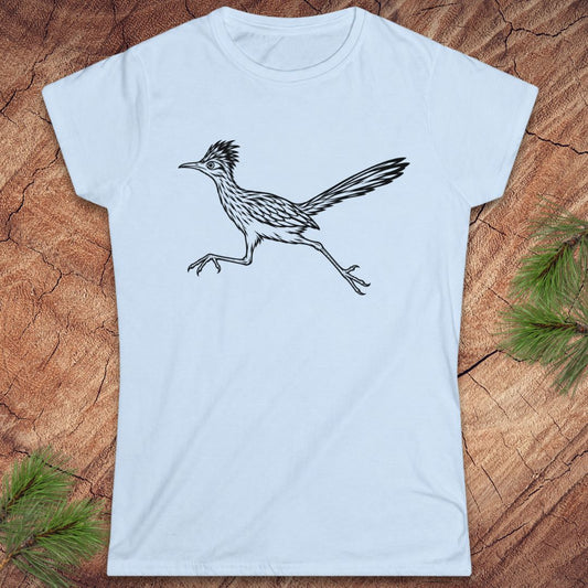 A light blue short-sleeve crewneck t-shirt featuring a black line art illustration of a running roadrunner bird, displayed flat on a rustic wood background with pine needles in the corners.