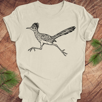 An off-white short-sleeve crewneck t-shirt featuring a black line art illustration of a roadrunner bird, laid flat on a rustic wood background with pine needles in the corners.
