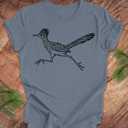 A slate blue short-sleeve t-shirt with a black line art illustration of a roadrunner, laid flat on a rustic wood background with pine needles in the corners.