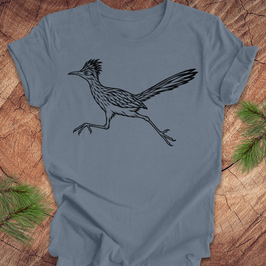 A slate blue short-sleeve t-shirt with a black line art illustration of a roadrunner, laid flat on a rustic wood background with pine needles in the corners.