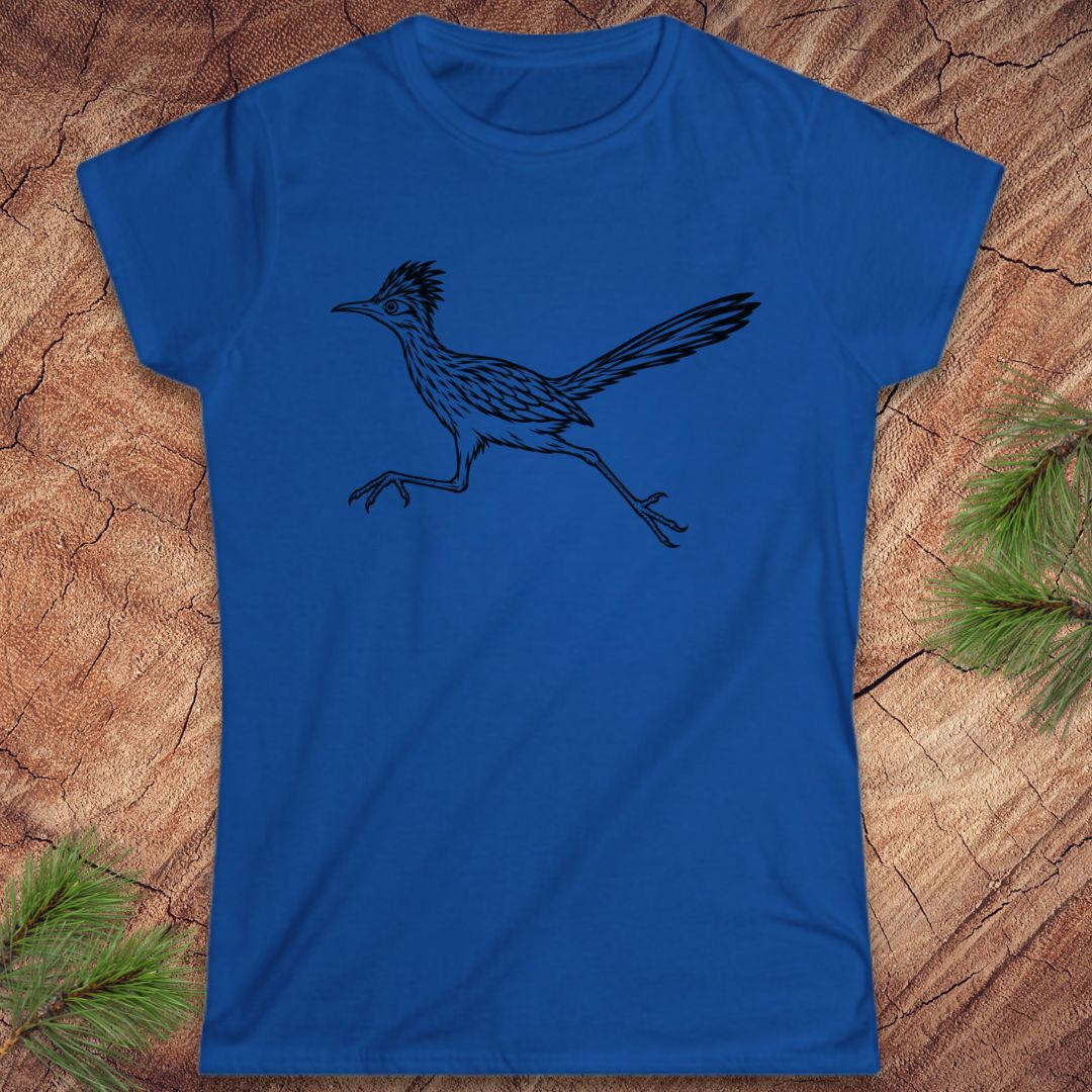A royal blue short-sleeve crewneck t-shirt featuring a black line art graphic of a running roadrunner, laid flat on a textured wood background with pine needles in the corners.