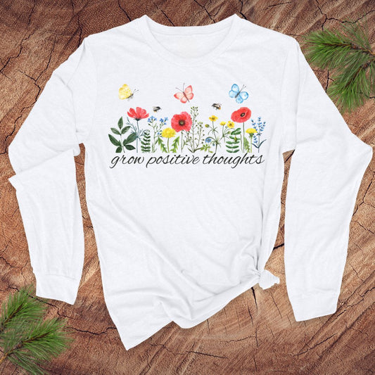 White long-sleeve shirt with floral design and "grow positive thoughts' text on a wooden surface