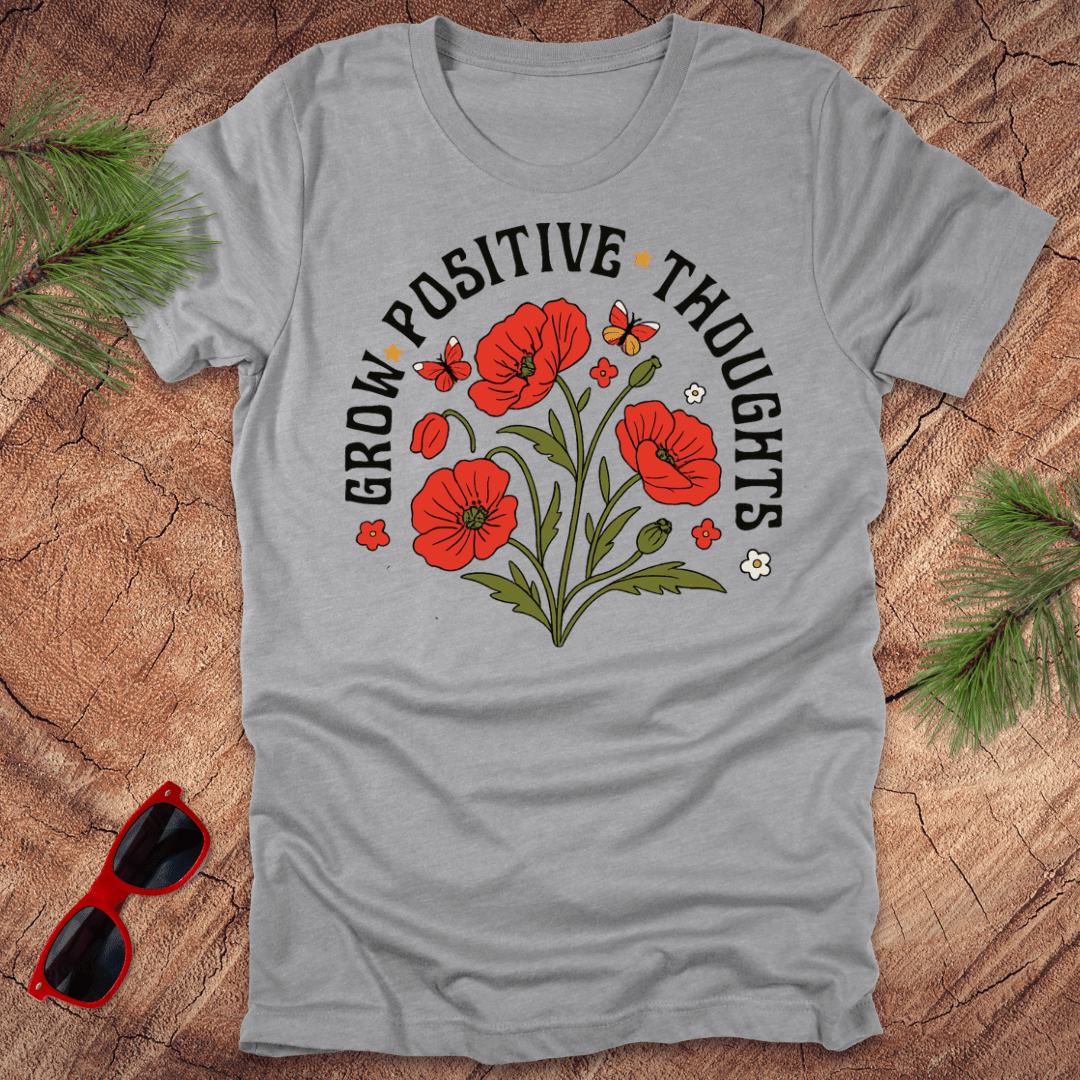 Gray t-shirt with red flowers and 'Grow Positive Thoughts' text on a wooden surface with sunglasses and greenery.