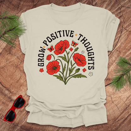 Beige t-shirt with red flowers and text on a wooden surface with sunglasses and greenery.