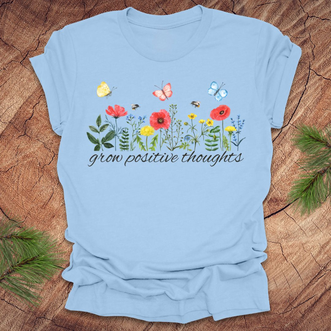 Light blue t-shirt with floral design and 'grow positive thoughts' text on a wooden surface.