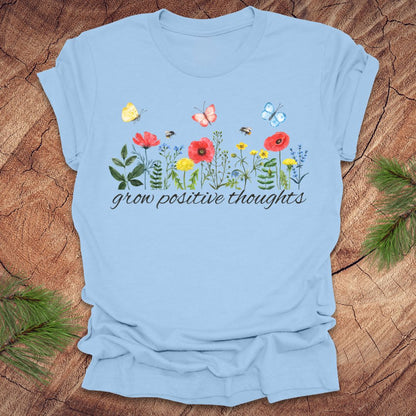 Light blue t-shirt with floral design and 'grow positive thoughts' text on a wooden surface.