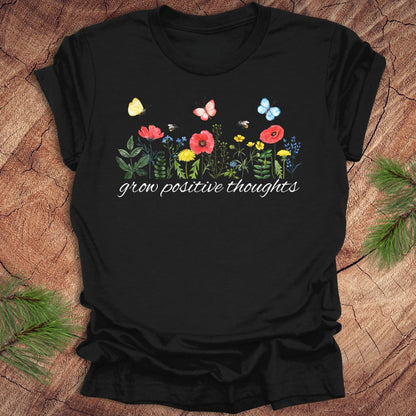 Black t-shirt with floral design and 'grow positive thoughts' text on a wooden surface.