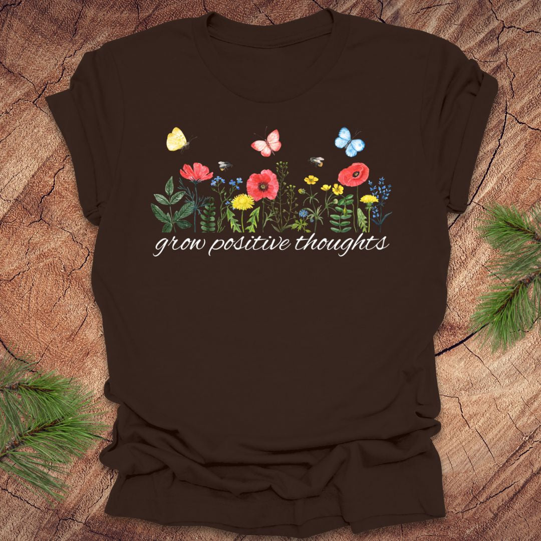 Brown t-shirt with floral design and 'grow positive thoughts' text on a wooden surface.