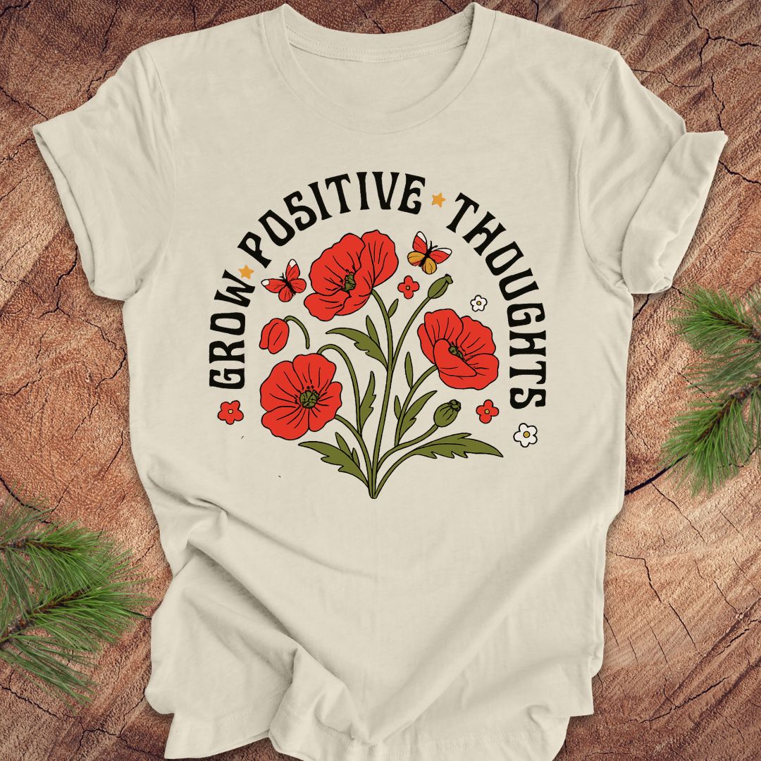Beige t-shirt with red floral design and text 'grow positive thoughts' on a wooden surface 