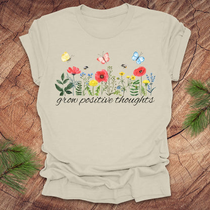 Beige t-shirt with floral design and 'grow positive thoughts' text on a wooden surface.