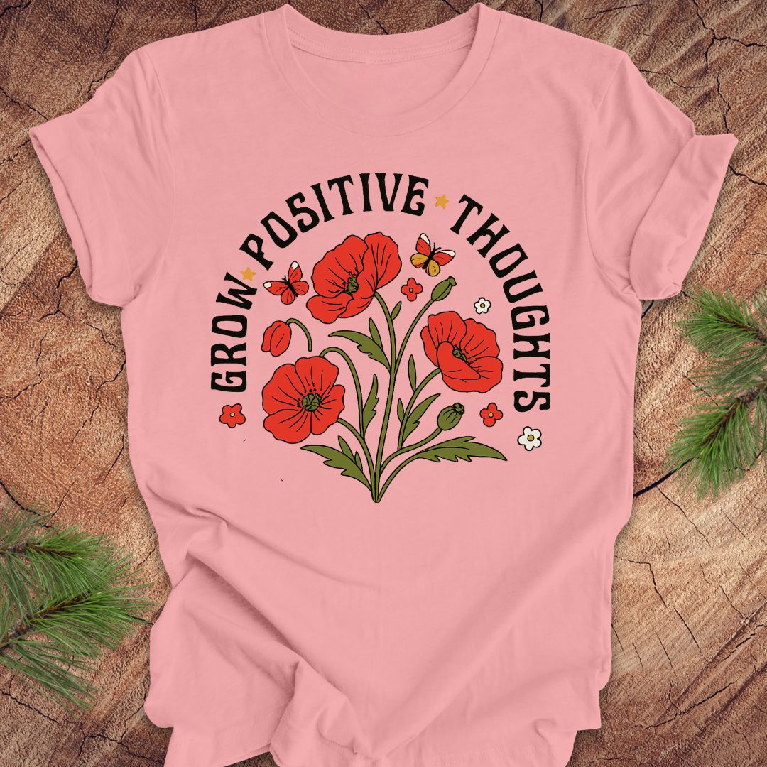Pink t-shirt with red floral design and text 'grow positive thoughts' on a wooden surface 