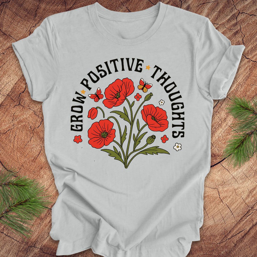Silver t-shirt with red floral design and text 'grow positive thoughts' on a wooden surface 