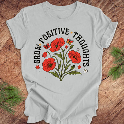 Silver t-shirt with red floral design and text 'grow positive thoughts' on a wooden surface 