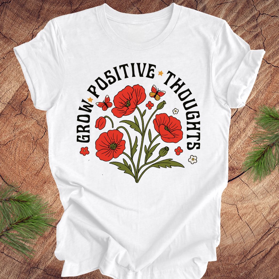 White t-shirt with red floral design and text 'grow positive thoughts' on a wooden surface 