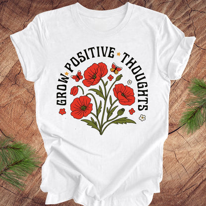 White t-shirt with red floral design and text 'grow positive thoughts' on a wooden surface 