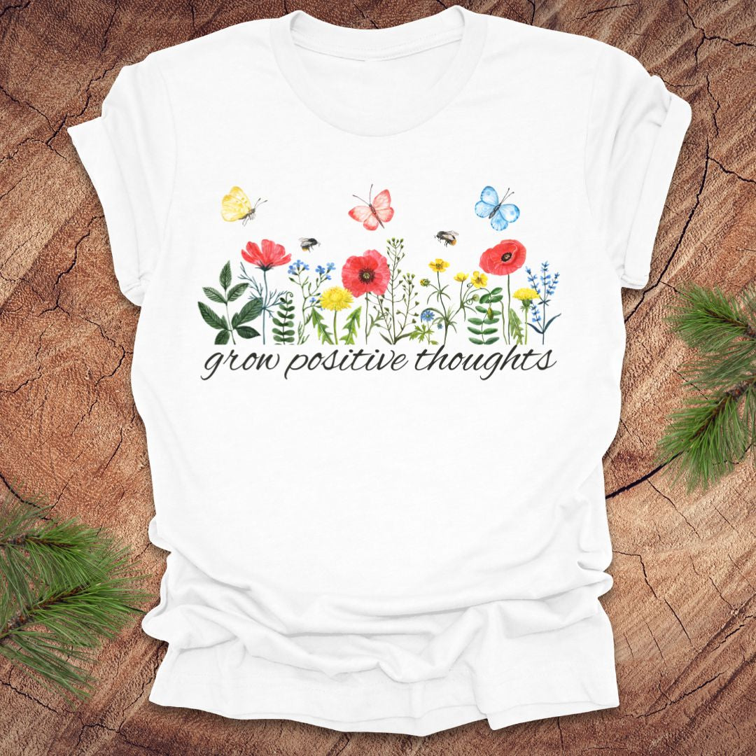 White t-shirt with floral design and 'grow positive thoughts' text on a wooden surface.