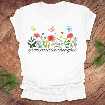 White t-shirt with floral design and 'grow positive thoughts' text on a wooden surface.