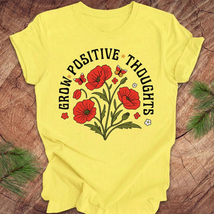 Yellow t-shirt with red floral design and text 'grow positive thoughts' on a wooden surface 
