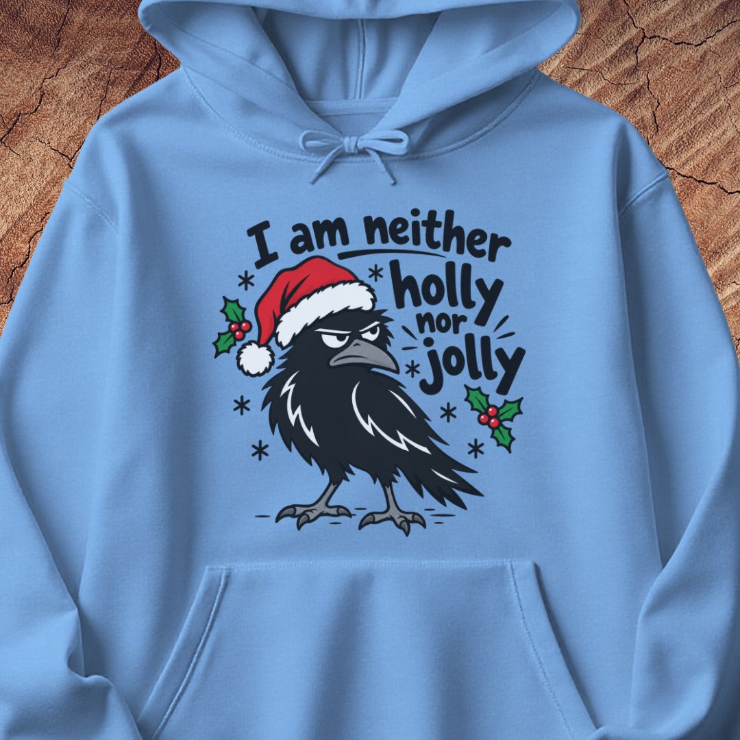 Carolina blue hoodie with a grumpy raven in a Santa hat surrounded by holly and snowflakes and the words I am neither holly nor jolly, laid flat on a rustic wood background.