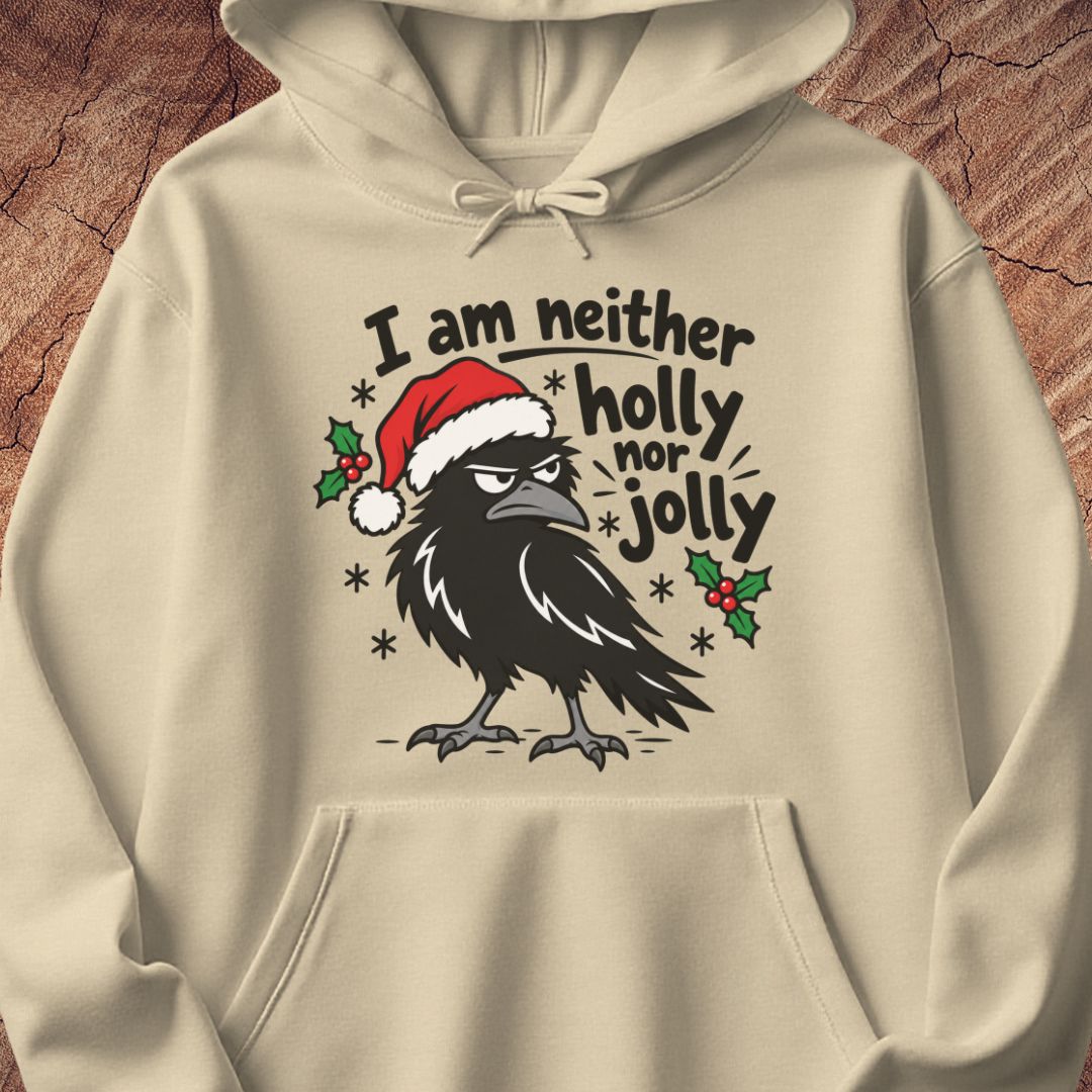 Sand beige hoodie with a grumpy raven in a Santa hat surrounded by holly and snowflakes and the words I am neither holly nor jolly, laid flat on a rustic wood background.